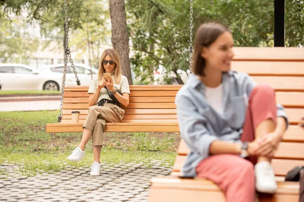 Genç, sarışın, günlük giysili, güneş gözlüklü, parkta ya da yaz günü meydanda salıncakta oturan ve akıllı telefondan kayan güzel bir kadın.