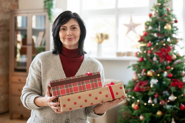 幸せな成熟したブルネット女性でカジュアルウェア保持スタックのギフトボックスで彼女の家族のためにプレゼントのために彼女の家族に対して装飾されたクリスマスツリー ホーム — ストック写真
