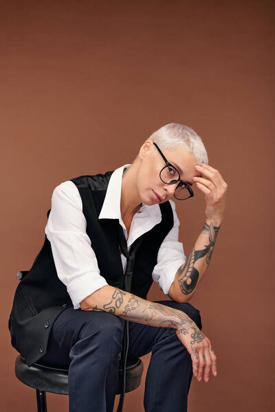 Pensive or stressed blond woman with arms covered with tattoos sitting on chair in front of camera while keeping hand on head in isolation