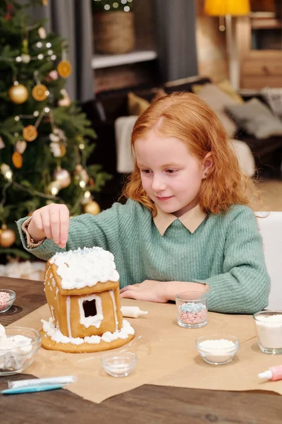 かわいい女の子はクリスマスパーティーのためのお祝いのデザートを準備しながらホイップクリームで飾られたジンジャーブレッドハウスの屋根を振りかける — ストック写真