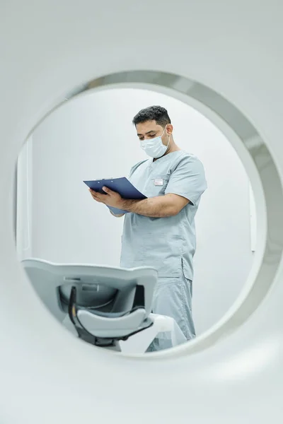 穿着制服 戴防护面具 站在超音速设备沙发旁在医疗文件上记笔记的成熟医生 — 图库照片