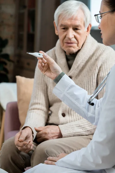 老年男子坐在年轻女医生旁边的沙发上 在测量了病人体温后看了看温度计 — 图库照片