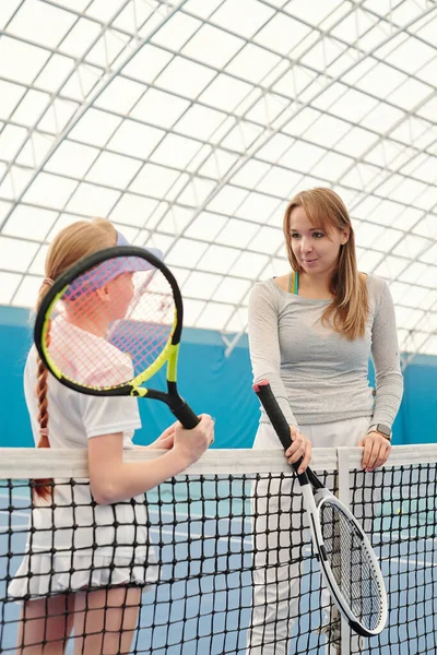 若いです女性テニストレーナーでアクティブウェアとかわいいです女の子とともにテニスラケットとともにネットでスタジアムでカメラの前に立って相互作用 — ストック写真