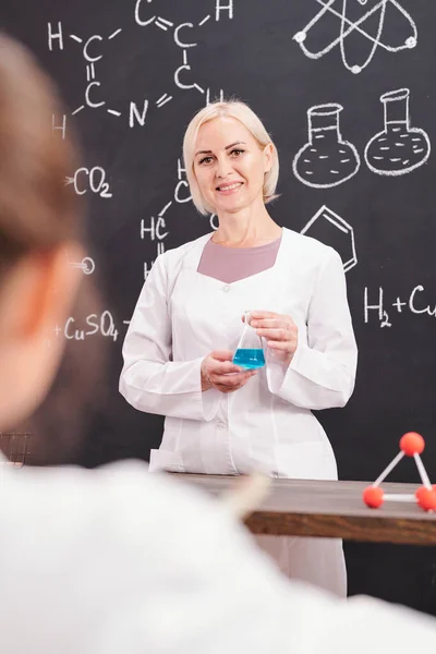 化学式で黒板に立っている間 青色液体物質を有するホワイトコート保持管中の化学の笑顔の先生 — ストック写真
