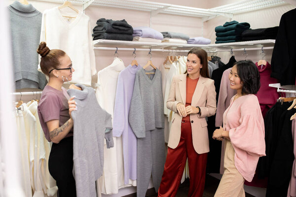 Happy young intercultural females standing in casualwear department during sale and looking at grey woolen sweater held by shop assistant