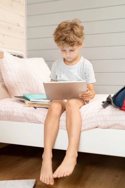 当代的小男孩 带着数码平板电脑坐在床上 在背包和书本中观看在线视频或课外电影 — 图库照片