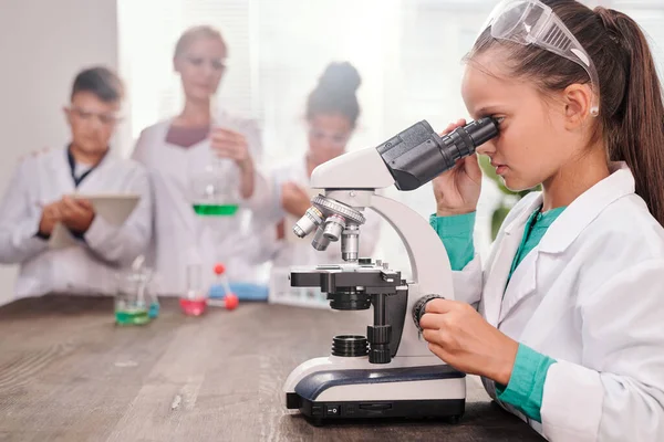 教室で化学実験を行う彼女のクラスメートと先生に対して机によって顕微鏡で見てホワイトコートの好奇心女子学生 — ストック写真