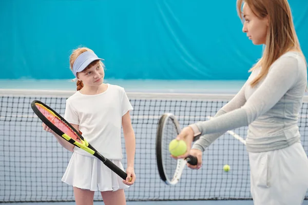 Active girl looking at female trainer and listening to her instructions about how to use ball and racket at the beginning of play on stadium