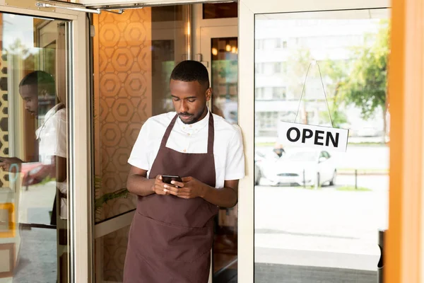 African young waiter in uniform standing in the door of cafe or resaurant and scrolling through new orders of clients in smartphone