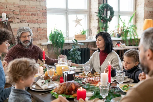 Three generations of large family sitting by served festive table and holding by hands while praying before Christmas dinner at home