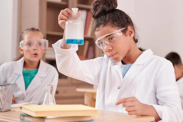 两个戴着防护眼镜的女学生和一个大嘴巴里看着蓝色液体物质的白人女孩在上课时被其中一个人抓住了 — 图库照片