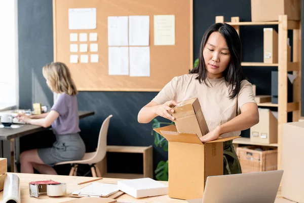 Çevrimiçi atölyede ya da depo odasında çalışan genç ve güzel bir işçi iş arkadaşına karşı dizüstü bilgisayar kullanma emri ile karton kutulardan birini paketliyor.