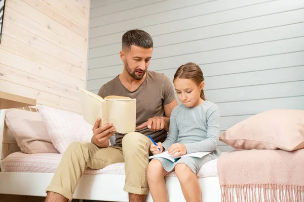 Küçük tatlı kızının notlarını gösteren açık bir kitabı olan genç bir baba. Hem yatakta oturuyor hem de ev ödevlerini yapıyor.