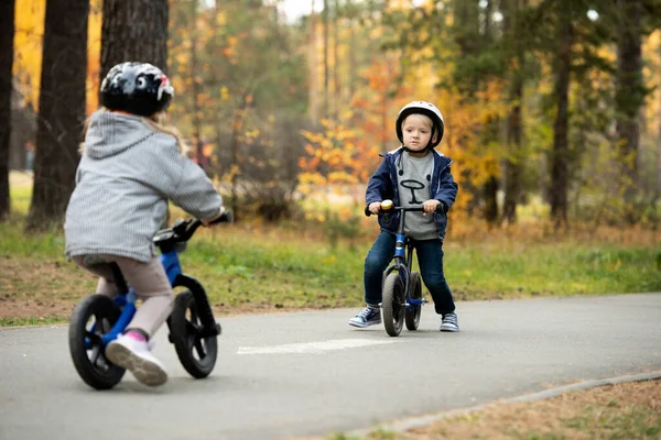 かわいいですブロンド男の子で安全ヘルメット見ます彼の友人移動上の彼に向かってバランス自転車で寒さの間に週末で自然環境 — ストック写真