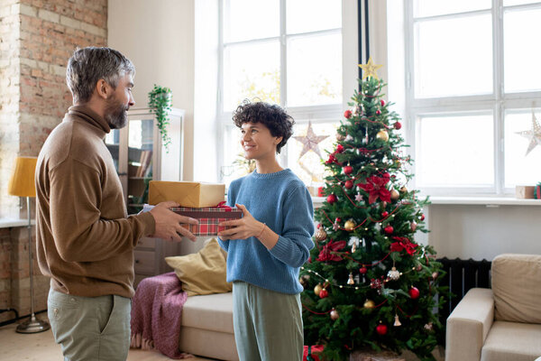 Young female with brown curly hair passing Christmas gifts to her husband and saying regards to him while both standing against decorated tree