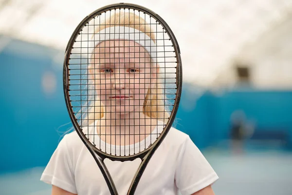小さなブロンド女の子でホワイトTシャツとヘッドバンド保持テニスラケットで正面と見ますあなたを通してネットで現代のスタジアム — ストック写真