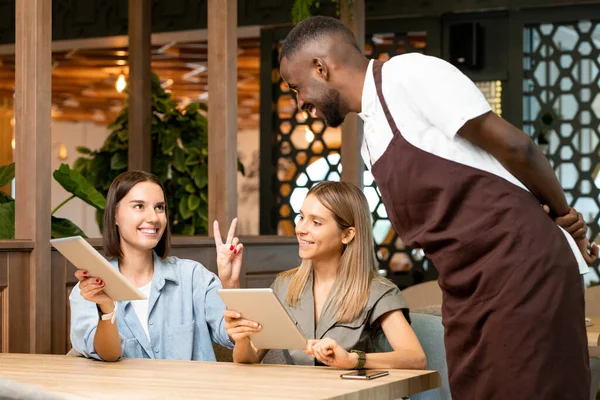 Üniformalı genç garson masanın üzerine eğilip tabletli iki kızdan birine bakıyor ve kafede online menüden bir şeyler sipariş ediyor.