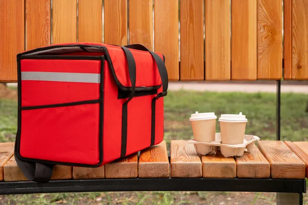 Big red bag with orders of clients of contemporary cafe or restaurant and two paper glasses with fresh hot coffee on wooden bench in park