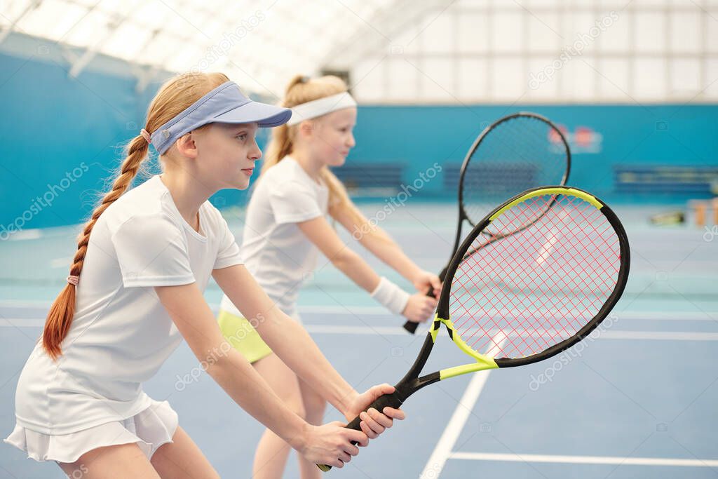 Two teenage girls in activewear standing on stadium with their legs bent in knees and holding tennis rackets ready to push the ball during play