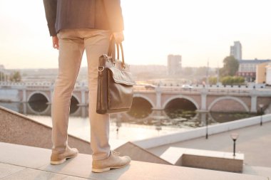 Nehir kenarındaki geniş merdivenlerin ve kentsel çevrenin önünde duran deri çantalı genç ve zarif iş adamının arka görüntüsü.