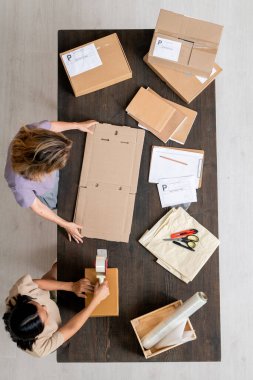 Geniş masa başında duran ve depolama odasında çalışırken müşterilerin siparişlerini kutulara koyan iki çevrimiçi mağaza yöneticisinin üst görüntüsü