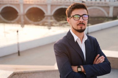 Takım elbiseli ve gözlüklü genç işadamı kollarını göğsüne kavuşturmuş kameranın önünde duruyor.