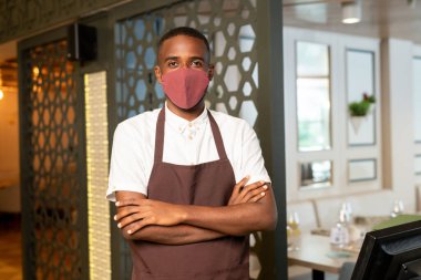 Modern kafe veya restoranda bilgisayar başında beklerken koruyucu maskeli ve kahverengi önlüklü çağdaş garson göğsünde çapraz kollar.
