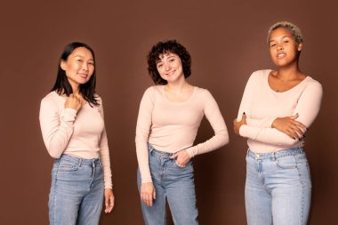 Young joyful women in white pullovers and blue jeans standing close to one another on brown background and looking at you with toothy smiles clipart