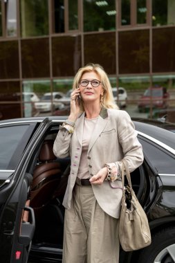 Zarif günlük giysiler içindeki ciddi sarışın iş kadını. Arabasının yanındaki cep telefonuyla dış bina karşısındaki iş ortağı.