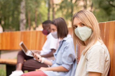 Koruyucu maskeli genç sarışın kadın akıllı telefon ve dizüstü bilgisayar kullanarak iki milenyum boyunca bankta otururken sana bakıyor.