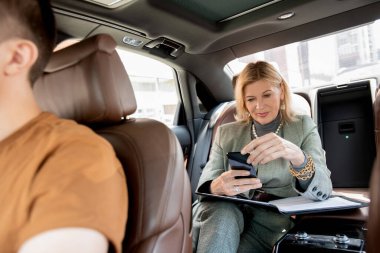 Sarışın, olgun bir kadın taksinin arka koltuğunda oturmuş iş görüşmesi için restorana giderken dokümanların üzerinden akıllı telefondan mesaj atıyor.