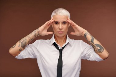 Kollarının her tarafında dövmeleri olan, erkeksi sarışın bir kadın. Kahverengi arka planda sana bakarken ellerini şakaklarına koyuyor.