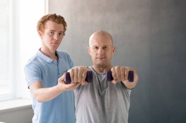 Mavi gömlekli profesyonel fizyoterapist ve elinde dambıllar olan yetişkin erkek hastası rehabilitasyon sırasında sana bakıyor.