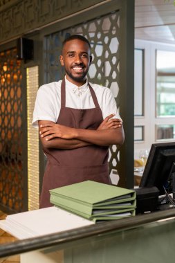 Young cheerful waiter of African ethnicity looking at you with toothy smile and crossing arms on chest while standing in restaurant environment clipart