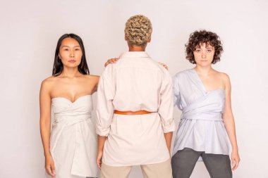 Genç Asyalı ve Kafkasyalı kadınlar ellerini beyaz giysili Afrikalı arkadaşlarının omuzlarında tutarken ikisi de sana bakıyor.