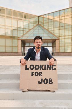 Resmi giysili genç sakallı bir adam elinde bir karton tutuyordu. Modern mimariye karşı merdivenlerde otururken iş aradığını söylüyordu.