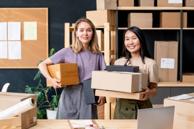 Çevrimiçi atölyede çalışan iki genç bayan ellerinde paketlenmiş siparişler, ellerinde kutular ve belgelerle rafların önünde duruyorlardı.