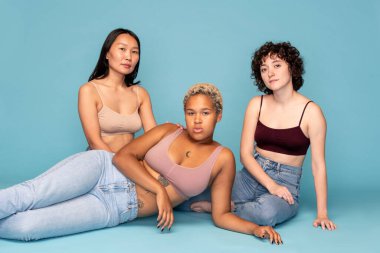 Üç çağdaş Asyalı, Kafkas ve Afrikalı kadın, mavi arka planda yerde dinleniyorlar.