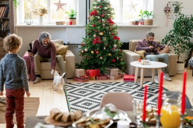 Akıllı telefonlu yaşlı bir adam köpekle oynarken oğlu yanında oturuyor ve cep telefonu ve torunu onlara bakıyor.