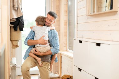 Mutlu, genç, sevecen bir baba, işten eve döndüğünde dizlerinin üzerinde evin koridorunda dikilirken küçük tatlı oğlunu kucaklıyor.