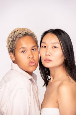 Afrikalı ve Asyalı etnik kökenli genç kadınların yüzleri, izolasyonda beyaz arka plana karşı kameranın önünde yan yana duruyorlar.