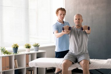 Profesyonel erkek fizyoterapist mavi gömlekli hasta destek kolu. Koltukta otururken halterlerle egzersiz yapıyor.
