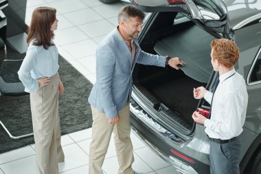 Çağdaş otomobil merkezinin genç yöneticisine danışıp araç seçerken yetişkin bir erkek müşteri yeni arabanın arkasında bekliyor.