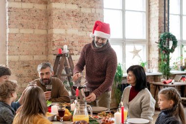 Noel Baba şapkalı olgun bir adam aile yemeği sırasında evde şenlik masasının yanında durup şarap kadehlerine ev yapımı içecek dolduruyor.