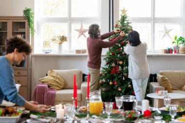 Olgun bir çift Noel ağacını koltukların arasında oyuncak toplarla süslerken, kız da ev yapımı yiyeceklerle şenlik masasına servis yapıyor.