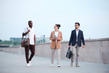 Kültürler arası üç genç iş ortağı akıllı günlük kıyafetleriyle nehir kenarına doğru ilerliyorlar ve tartışıyorlar.