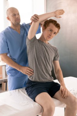 Rehabilitasyon kliniklerindeki yan bükme egzersizi sırasında tıbbi kanepede oturan, üniformalı, kel, olgun bir fizyoterapist.