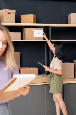 Paketlenmiş karton kutudan sipariş içeriği hakkında bilgi okuyan koyu saçlı genç bayan yöneticinin yan görünümü