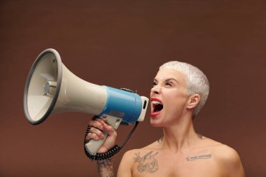 Kolunda, göğsünde ve boynunda dövmeleri olan, megafonla bağıran ve önemli bir duyuru yapan çok kızgın sarışın erkek gibi bir kadın.