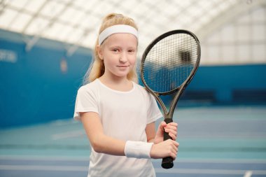 Cute blond little girl in white activewear holding tennis racket by left shoulder while standing in front of camera in stadium environment clipart